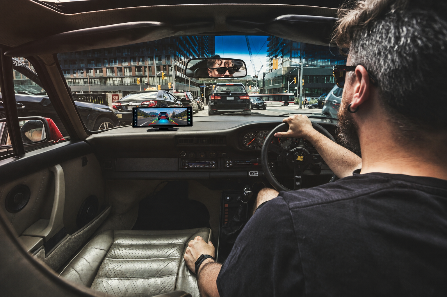 A man wearing sunglasses sits in the driver's seat of a vintage car, looking out through the windshield at a city street scene. The car interior features a beige leather seat and dashboard, and a small rectangular screen is mounted on the dashboard.